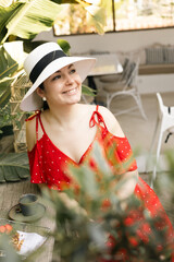 Smiling woman in red dress and sunhat enjoying morning coffee in tropical garden cafe setting