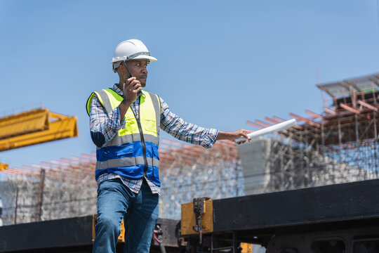 Engineer with Plans and Radio at Building Site, African American Foreman Directing Work on Construction Project, Construction Site Manager Communicating on Walkie-Talkie - Powered by Adobe
