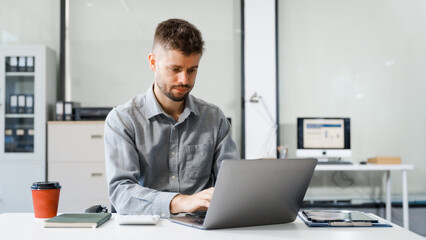 Fototapeta premium A smart Caucasian businessman sits confidently at desk in modern indoor office, using laptop for online consulting, showcasing professionalism, success, digital expertise in corporate environment.