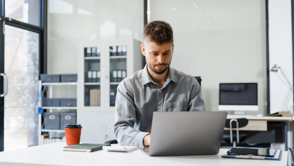 A smart Caucasian businessman sits confidently at desk in modern indoor office, using laptop for online consulting, showcasing professionalism, success, digital expertise in corporate environment.