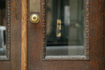 A detailed close-up shows a brass doorknob on a dark wooden door with carved decorative trim. Glass panels reflect the building across the street, adding depth to the composition.