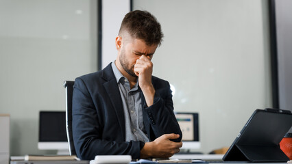 A stressed Caucasian businessman in a suit sit indoors at an office desk, suffering from office...