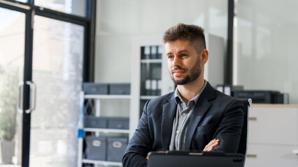 Fototapeta premium A smart Caucasian businessman sits confidently at desk in modern indoor office, using laptop for online consulting, showcasing professionalism, success, digital expertise in corporate environment.