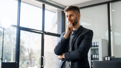 A smart Caucasian businessman sits confidently at desk in modern indoor office, using laptop for...