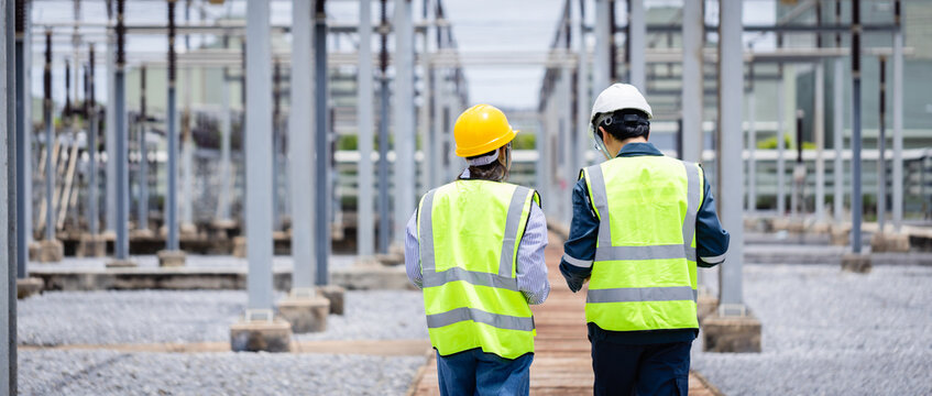 Engineer is working at high voltage power substation wearing safety gear while conducting inspection and check of electrical infrastructure to ensure proper operation and safety