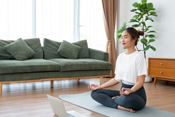 Young Asian woman meditating in lotus position at home, sitting on the floor in fitness clothing. Breathing workout to achieve relaxation, peace and mindfulness
