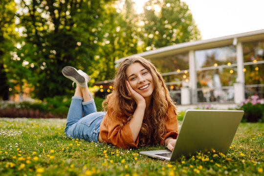 Portrait of a cute woman with a laptop relaxing on the grass in the park. Smiling female freelancer enjoying work outdoors. Remote work and nature concept.