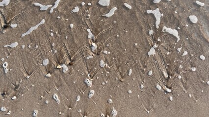 A sandy beach with intricate wave patterns formed by receding water. The foam and bubbles add texture and contrast to the smooth surface, creating a visually appealing coastal scene