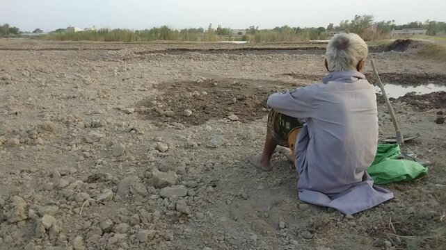 Exhausted elderly farmer resting alone on dry barren land after hard work, gazing silently at the lifeless earth


