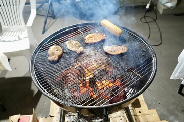 Close-up view of various meats and corn on the cob grilling over hot coals on a charcoal barbecue, with smoke rising. An outdoor summer cookout, party, or casual dining experience concept.