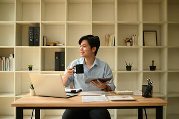 An Asian professional man smiles while holding a coffee mug and tablet, enjoying a relaxed moment during his workday at a modern, organized office desk