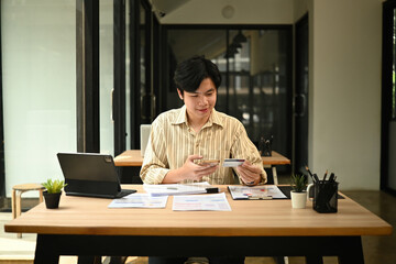 Young Asian man makes an online payment using a smartphone and credit card at a modern office desk