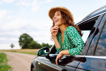 Traveler in hat and bright dress leaning out of car window. Beautiful woman in hat enjoying car...