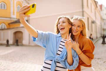 Two cheerful women having fun and taking selfies on their phones on sunny streets. Beautiful female...