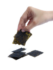 A hand picking up a square piece of dried seaweed snack with a stack of seaweed sheets on a white background, isolated for commercial food presentation or packaging use.