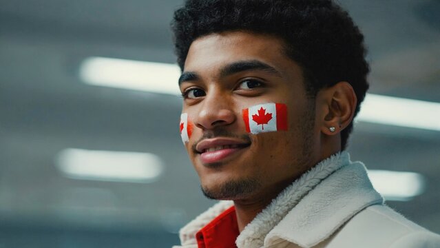 Young Black man with curly hair and Canadian flag face paint smiles confidently. Indoor setting with bright lights. Celebrating Canadian culture.