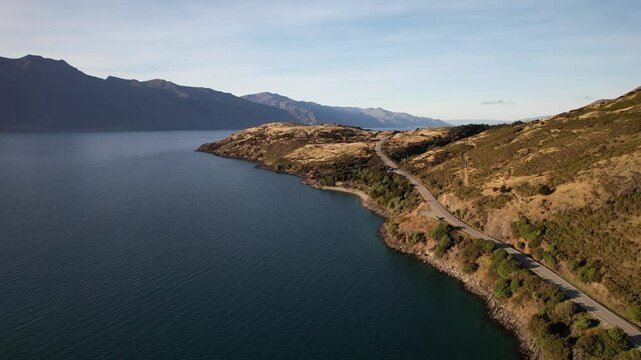 Coastal Road Along Golden Hills On Shore Of Lake Hawea In Otago Region, New Zealand. Mountain Range In Background. aerial pullback shot