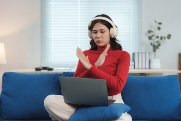 Young woman wearing headphones showing stop sign with crossed arms while using laptop on sofa