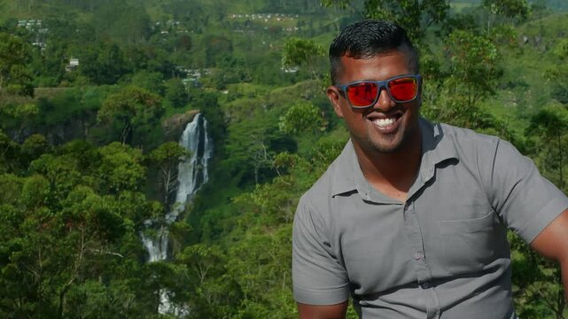 Cheerful young male wearing vibrant red sunglasses sitting near stunning Ramboda Falls, capturing scenic tropical landscape of Sri Lanka with genuine joy and wanderlust spirit