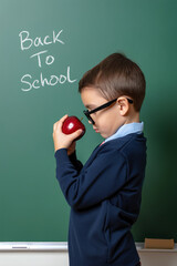 A boy in a navy sweater stands in front of a chalkboard, holding a shiny red apple with intent. The words back to school are scribbled behind him. Back to school concept. Generative AI.