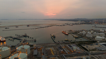 large industrial oil and gas storage tanks at a coastal refinery terminal during dusk. Tanker ships docked at long piers highlight the global energy transport infrastructure.