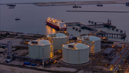 Aerial view of an LNG tanker docked at an offshore terminal with multiple pipelines, storage tanks, and an industrial complex. Captures global energy logistics