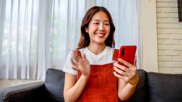 Smiling young asian woman video calling at Living Room, Waving Hello into Smartphone Camera. Talking Online With Friend, Happy lady talking by virtual chat meeting dating online using mobile videocall
