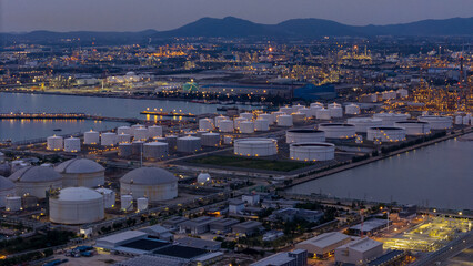 large industrial oil and gas storage tanks at a coastal refinery terminal during dusk. Tanker ships docked at long piers highlight the global energy transport infrastructure.