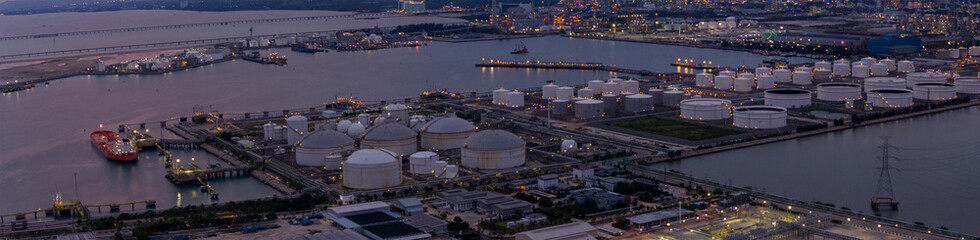 large industrial oil and gas storage tanks at a coastal refinery terminal during dusk. Tanker ships docked at long piers highlight the global energy transport infrastructure.