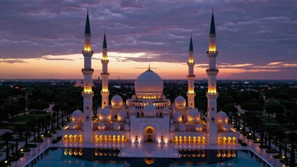 Aerial video captures a grand mosque at sunset, showcasing its illuminated domes and minarets reflected in a tranquil pool, with a dramatic sky overhead. - Powered by Adobe