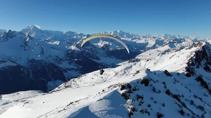 Paraglider soars above snowy mountain peaks under a clear blue sky on a bright sunny winter day
