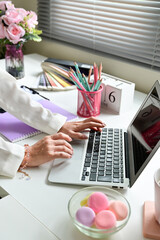 A woman is typing on a laptop in front of a white desk. The desk is cluttered with various items...