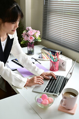A woman is typing on a laptop in front of a white desk. The desk is cluttered with various items...