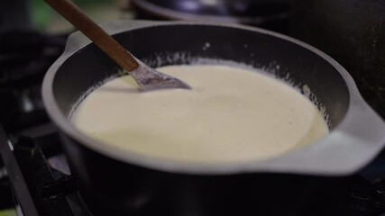 Creamy Bagna Cauda sauce simmers gently in a black pan as it is stirred with a wooden spoon, blending anchovies, garlic, butter, and olive oil in a warm Piedmontese kitchen setting.