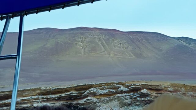 Distant establishing overview of Candelabra geoglyph on sandy Paracas hillside beneath cloudy desert sky, from tour boat