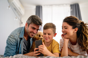 Happy parents with kid using smartphone at home together, playing, having fun with dog