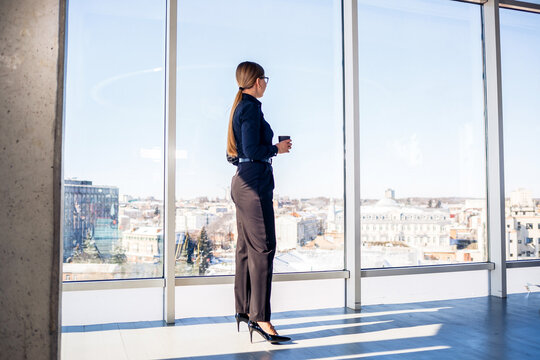 A determined businesswoman in her sleek corporate office is gazing out the expansive window while holding a steaming cup of coffee, deeply contemplating her next significant career move