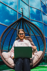 A modern professional woman works remotely in a hanging chair outside a contemporary building,...