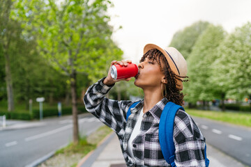 Young latin student drinking a refreshing soda from a can while walking on the street next to a park