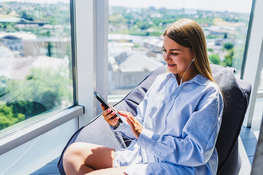 A young woman is leisurely relaxing by a window, using her smartphone and wearing headphones, completely absorbed in the music that brings her enjoyment in a stylish and modern setting