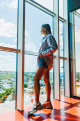 A woman stands at a window, gazing at the modern urban landscape with stunning architecture, dressed in stylish clothing, illuminated by soft daylight that enhances the serene atmosphere