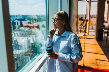 A contemplative woman is savoring a scenic view through a large window while holding a cup of coffee in her hand at a modern office space, perfectly embodying tranquility and inspiration
