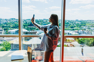 A young woman is happily taking a selfie with her smartphone in front of a scenic urban view seen through a large, modern window in a bright, airy space filled with daylight and warmth