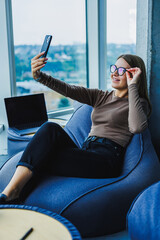 A young woman is savoring a peaceful moment as she takes a stylish selfie in a cozy environment, all while enjoying stunning views of the city. This upbeat moment captures happiness and leisure