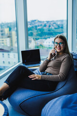 A dedicated young woman in a stylish outfit works hard in her beautifully decorated workspace, using modern technology and enjoying a stunning view of the vibrant cityscape through a large window