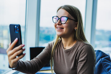 A young woman is on a lively video call in a modern workspace, smiling while using her smartphone. She radiates cheerfulness and professionalism as she connects with friends or colleagues