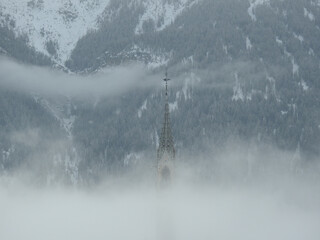 Zwischen Nebelschwaden ein Kirchturm im Stil der Neogotik, dahinter die drohenden Berghänge der...