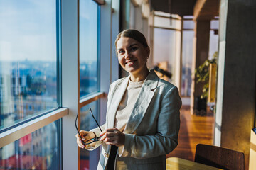 In a modern office setting that offers a stunning view of the city, a professional woman stands confidently while holding her glasses with a warm smile, embodying the essence of success and ambition