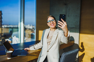 A confident and professional woman happily taking a selfie in a modern office setting that boasts a stunning city view, reflecting her successful career and trendy lifestyle in the business world