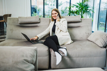 A professional woman is working from home on her laptop, engaged in conversation while sitting comfortably on a cozy sofa in her modern living space, blending productivity with relaxation